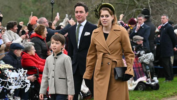 Princess Beatrice wearing a camel coat and green hat walking towards the church with her husband wearing a dark suit with matching green tie accompanied by his son wearing a grey jacket with green collar.