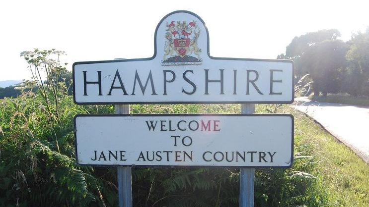 A white road sign saying "Hampshire - Welcome to Jane Austen County". It's on the side of a country road with ferns and other greenery behind it. It's a sunny day.