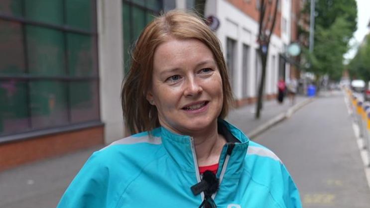 Anne Madden has short ginger hair and is smiling at the camera as she looks to her left. She has a blue coat on, and behind her is brown and white buildings.