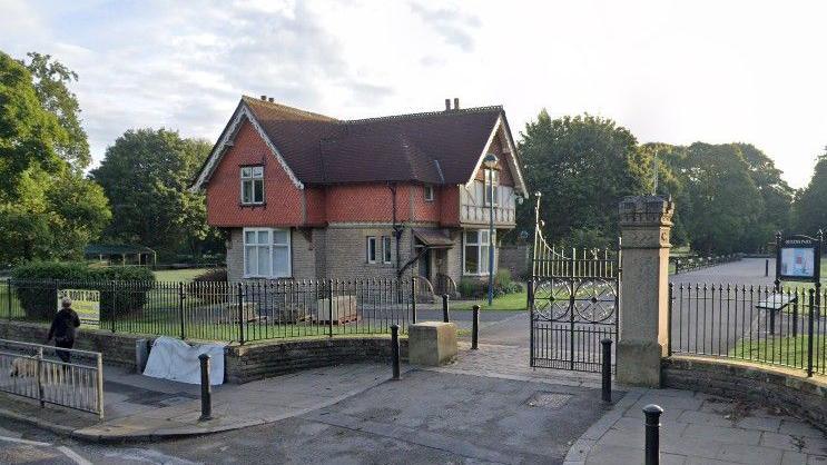 Park railings outside entrance with old two-storey house behind the gates. Trees can be seen in the background.