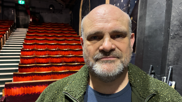 Peter Beck is off centre to the right looking at the camera. He has a short grey beard and is wearing a blue T-shirt and green jacket. He is stood in front of a theatre's seating.