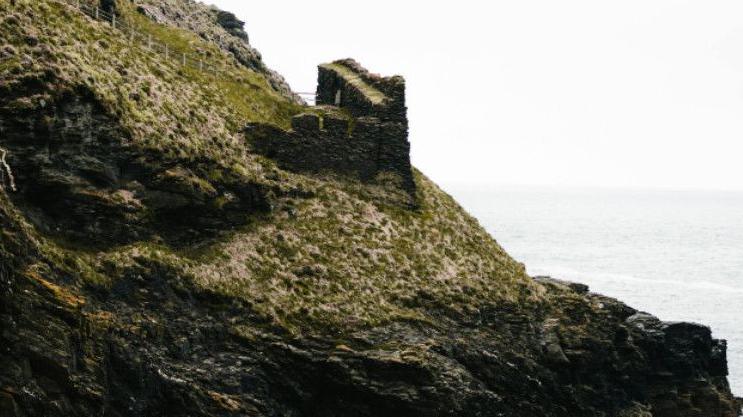 A cliff side with remains of an old industrial building, set against a backdrop of the sea and a grey sky