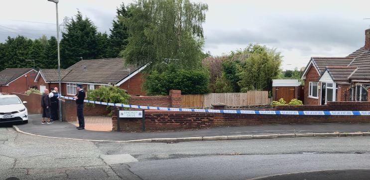 Police tape cordons off an area including Finchley Drive around Chadwick Road with two police officers in attendance as a resident in the street looks on.