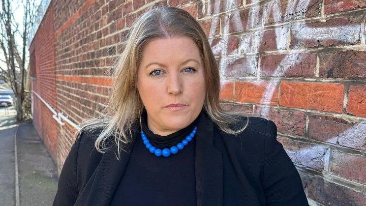 Donna Jones standing in an alleyway which has one piece of graffiti on the wall. She is looking serious and is dressed in black with a string of large blue beads around her neck