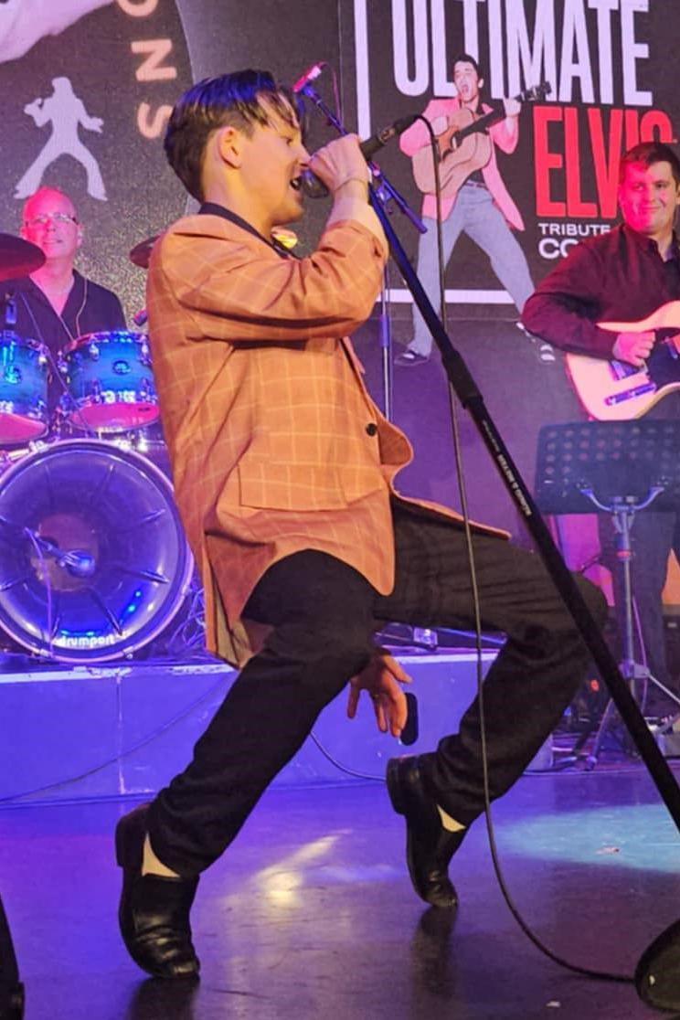 Cooper is singing on stage. He is leant back with bent knees and he is standing on his toes in a typical Elvis-style dance move.