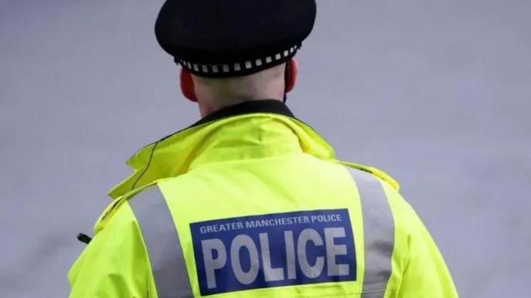 Police officer in GMP jacket faces away from camera. He is wearing a neon yellow jacket which has Greater Manchester Police written on a blue background with grey stripes down either side. He is wearing a police hat. 