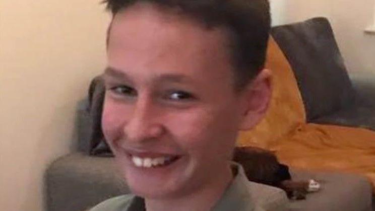 A young boy smiles whilst looking at the camera. He appears to be in a living room. He is wearing a khaki coloured shirt over a white T-shirt.