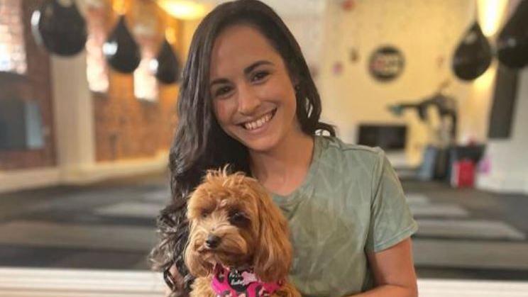 Leanne Lucas, with long, dark and curled hair, smiling at the camera with a cockapoo-type dog on her lap