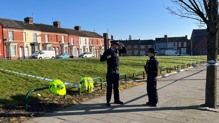 Two police officers stand by a cordon in front of a small grassed area near terraced houses
