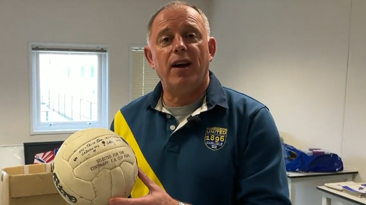 Simon Clarkson, who has short grey/brown hair and is wearing a blue and yellow Carlisle United top, is holding up an old football which is signed in pen.