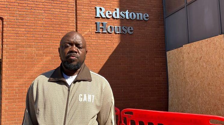 A man with short dark hair and a beard, wearing a beige zip-up jacket. He is standing in front of a brick-built block of flats with several storeys, with a glass entrance porch and "Redstone House" in silver lettering on a brick wall. There is wooden boarding around the entrance porch, and a low red temporary fence in front of the entrance.