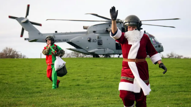 A man dress as Father Christmas in a red suit waving. He is stood in front of a large grey military helicopter. There is a man dressed as an elf in green and red holding a sack of presents behind him