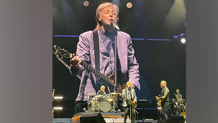 Sir Paul McCartney wearing a grey blazer, stands in front of a microphone, holding a guitar, on a huge stage. Bruce Springsteen is to his left, smiling at him, as he holds a guitar. A man is on the drums behind them.