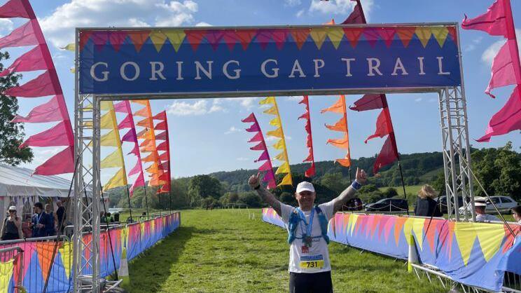 A man wearing a white cap and a white shirt stands at the finish line of a race with his thumbs up. There are multi-coloured flags around him. A banner above his head reads "Goring Gap Trail".