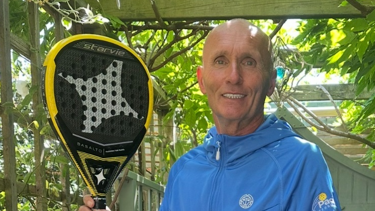 Alan Jordan is standing to the right of the image and is wearing a blue jacket and smiling. He is holding a black and yellow raquet.