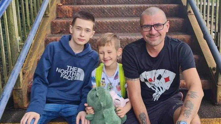 Jaroslaw Rossa and his sons Filip and Dominic - Jaroslaw is wearing glasses and a black T-shirt with a design showing playing cards on it. Dominic is sitting in the middle holding a stuffed toy and Filip is wearing jeans and a blue top. They are sitting on steps at a train station.