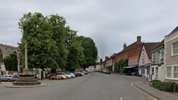 Dedham's High Street with shops, houses and pubs on the right and a war memorial and trees on the left. Cars are parked either side of the road, with the parish church just about visible behind the trees.