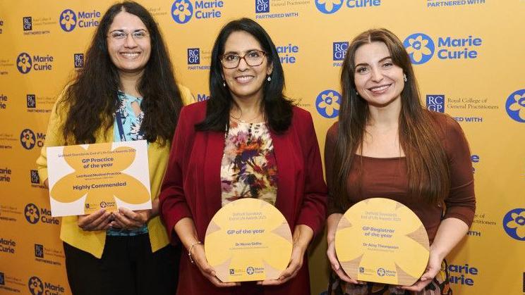Three women stand in front of a yellow board saying 'award winners' and 'Marie Curie' on it. They all hold a yellow certificate.