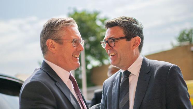 Sir Keir Starmer and Andy Burnham shaking hands and smiling broadly at each other. Sir Keir has short, grey hair and Burnham has short, dark hair. They are both wearing glasses and grey suits with white shirts. Sir Keir is wearing a brown/maroon tie and Burnham a grey tie.
