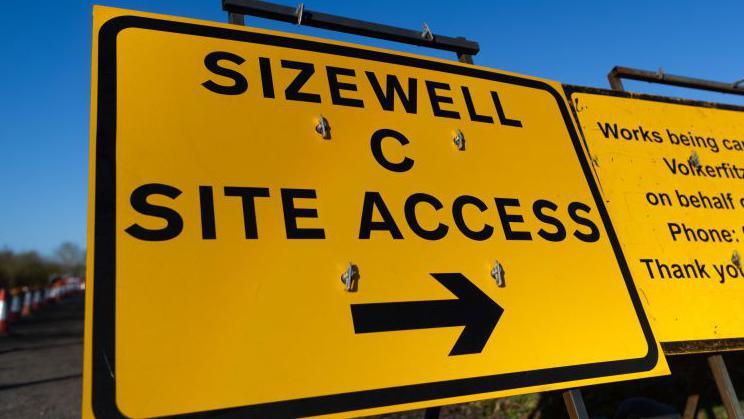 A yellow sign that reads in black writing: "Sizewell C site access." A black arrow to the right directs visitors which way to go. Another yellow sign with black writing off picture is next to this sign.