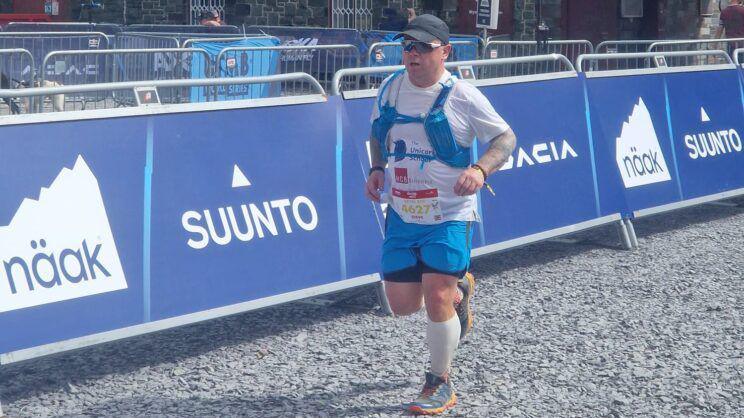 A man wearing blue shorts and long white socks runs on a gravel path. There are blue barriers to one side of him. He has a race number attached to his top.