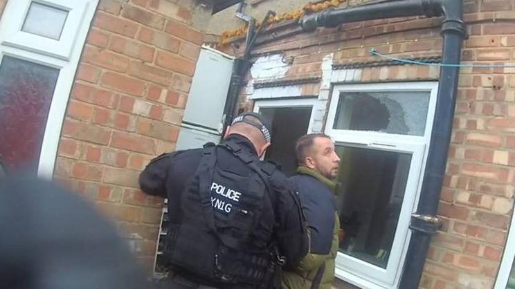 Body worn camera footage of a man with a green and black puffer coat on standing outside a property, with a police officer wearing a black uniform and hat. The police officer looks like he is arresting the man.