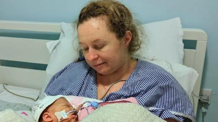 Katie Fowler sits in a hospital bed and is holding her baby Abigail. Abigail has a tube in her mouth and a pale hat on her head, while Katie is wearing a striped shirt and looking down at her baby.