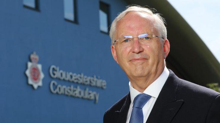 PCC Chris Nelson wearing a black blazer, white shirt, and blue tie. He has white hair with a side parting on the left, and clear framed glasses. He is standing in front of the Gloucestershire Police headquarters, looking at the camera. The Gloucestershire Constabulary sign and logo is visible on the building behind him. 