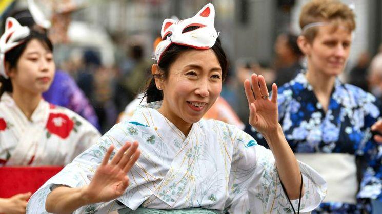 A woman in the foreground dressed in a white patterned kimono with a cat mask on top of her head