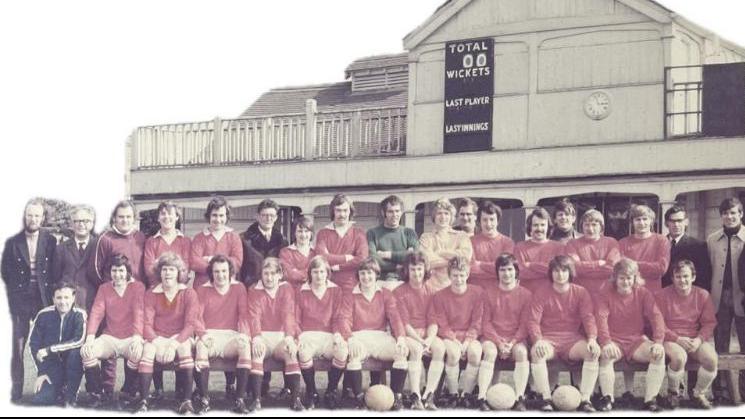 A football team dressed in red shirts and white shorts line up for a photograph in two rows, one sitting and one standing