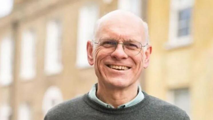 Tim Bick smiling and wearing a grey jumper and a blue shirt underneath