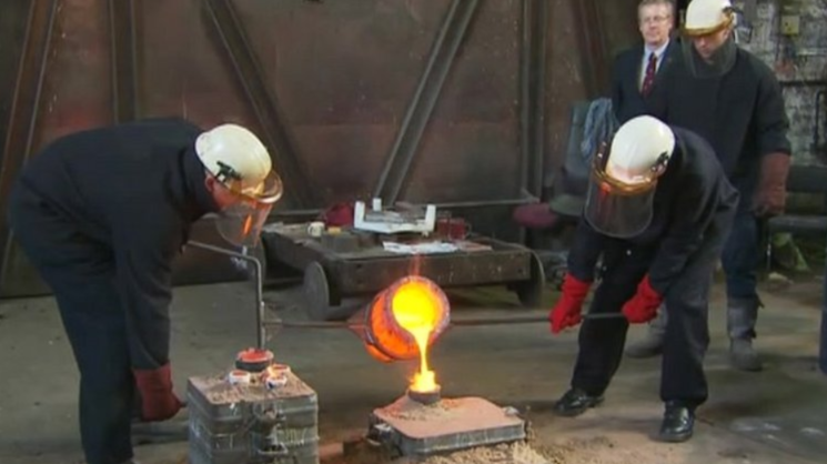 Two men in protective clothing pour molten metal into a bell cast