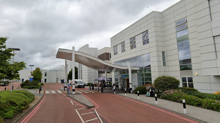 The front entrance of a hospital. The building is large and white with lots of windows. There is a pedestrian crossing over a road which leads to the entrance. Over the entrance and part of the crossing is a large wave-shaped roof awning.
