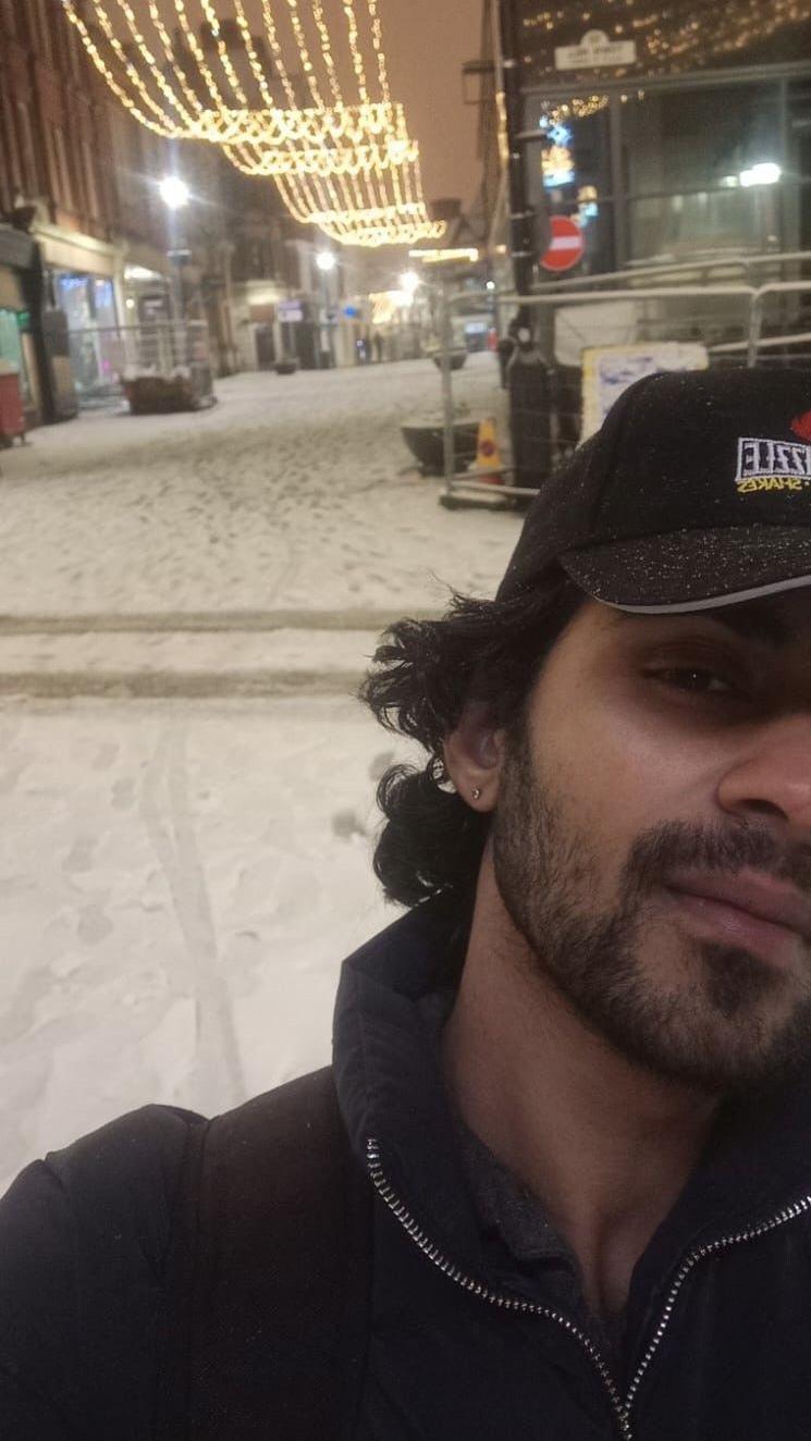 A young Indian man witha  slight beard and wearing a baseball cap and black zip-up jacket, taking a selfie in a snowy city street with Christmas lights in the background