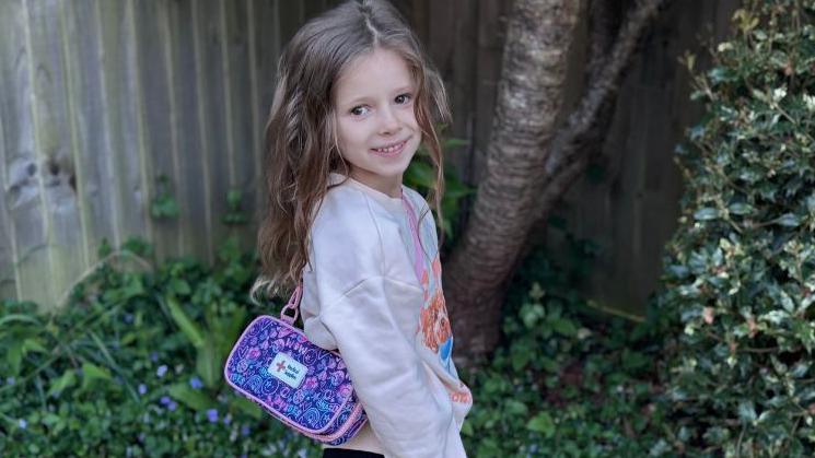 Margo standing in the garden, with her epipen case over her shoulder, which is purple and pink in colour. There are bushes around her and she has on a beige jumper and dark trousers