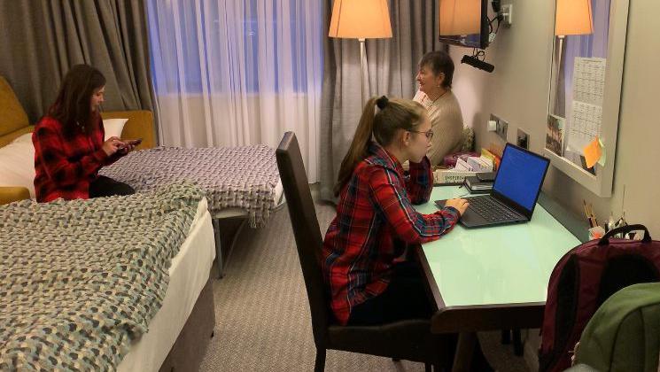 Kateryna yn gwneud gwaith cartref wrth fwrdd mewn ystafell gwesty, tra bod ei mam yn eistedd ar wely a'i nain mewn cadair wrth ei hymyl