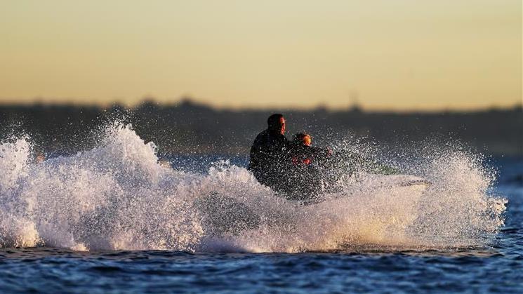 A sunset on the river with one adult and a child are on a personal water craft. Water is spraying up as the water craft is turning. 