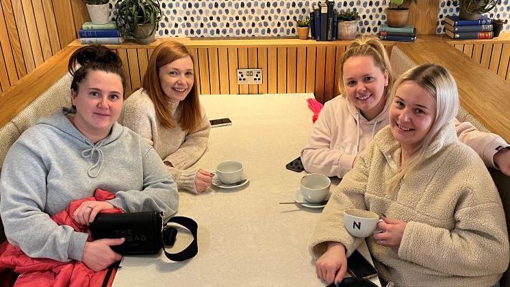 Four young women are sitting around a table in a cafe booth. They each have a cup and saucer.