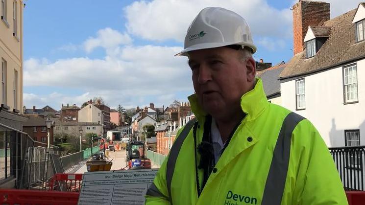 Stuart Hughes wearing a high visibility jacket and hard hat, he is stood on the bridge. 