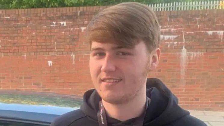 Ellis is standing in front on a red brick wall. He has short hair and is wearing black hoodie with a lanyard around his neck.