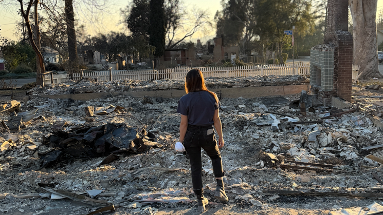 A woman standing in the middle of debris from the wildfires. In the background are trees and a fence. To the right is what looks like the bricks from a fireplace.