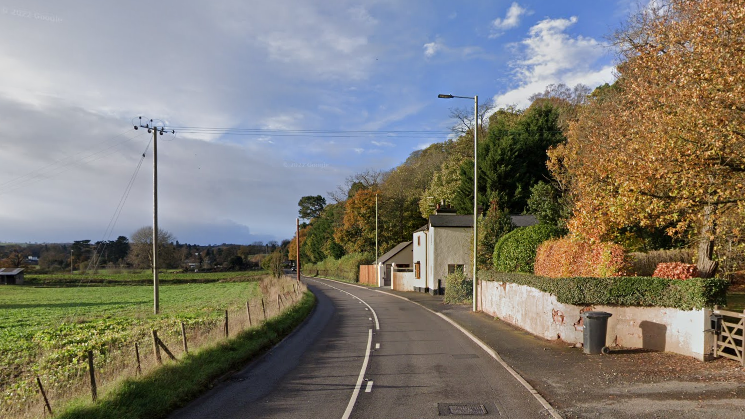 A long road curving around a bend, with a white house on the right-hand side. On the left, separated by a small fence is a green field. There are trees behind the house and along the right-hand side of the road