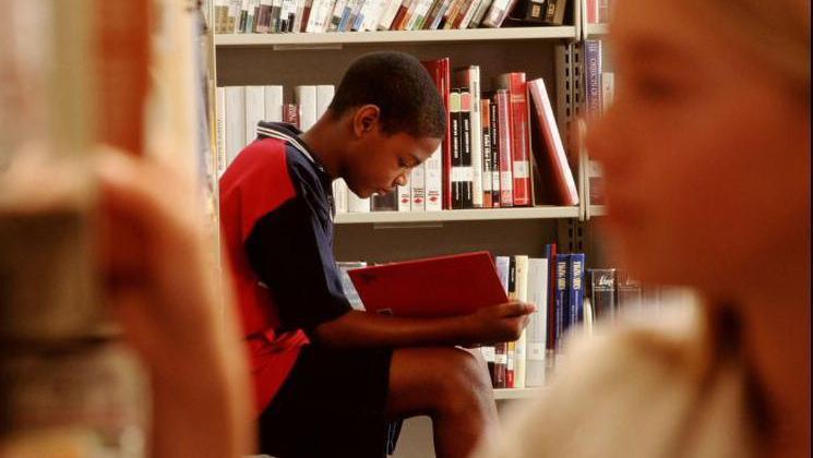 A boy reads in a library, while a girl picks a book from a shelf