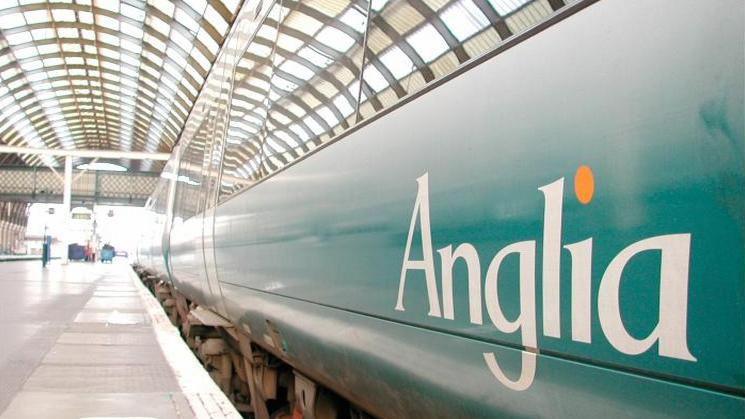 An Anglia Railways Class 170 train, bearing its white logo with orange dot above the 'i', on a turquoise background. It is standing within a station with arched glazed roof and a footbridge can be seen in the distance.