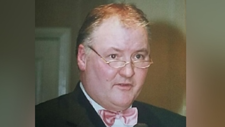 A man in a pink bow tie, with white shirt and black dinner jacket. he is wearing half moon spectacles with gold rims and appears to be in the middle of speaking. The tip of a microphone can just be seen at the bottom of the picture. He is standing in a room with cream walls.