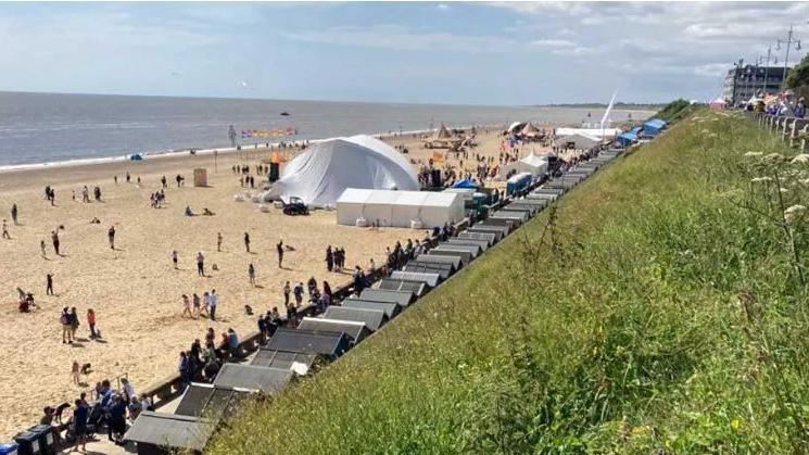 A white tented dome on a beach with lots of people gathered around. There is grass in the foreground of the photo.