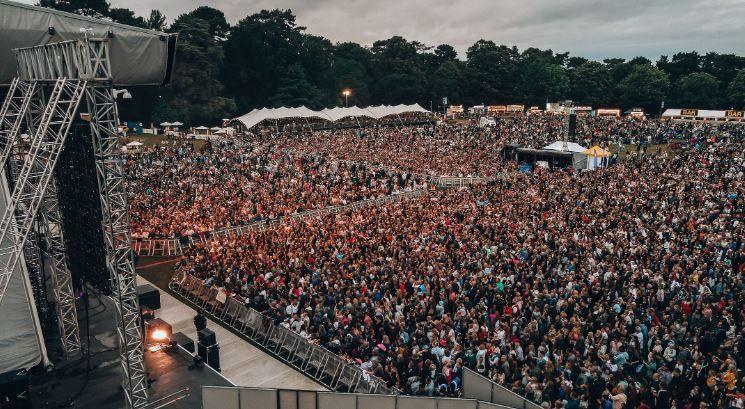 An outdoor festival audience, pictured from one side of the stage, showing the mixing desk area and tents/foodstalls around the arena perimeter.