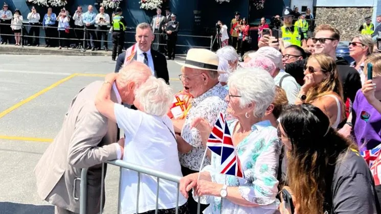 The King being kissed on cheek by Guernsey resident Kathleen Moriarty, at the front of a large crowd on a sunny day.