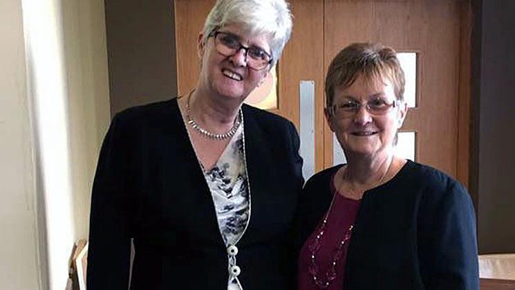 Two women stand side by side. The taller woman on the left has grey short hair and is wearing glasses and a black cardigan and white and blue top. The woman one right has short dark hair, glasses and is wearing a burgundy top and blue cardigan. Both are smiling at the camera. 