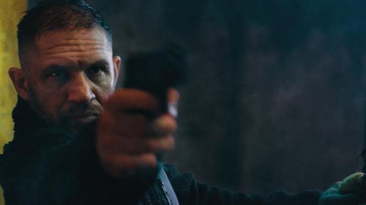 Tom Hardy in character, pointing a gun at the camera. He has short dark hair and is wearing all black.